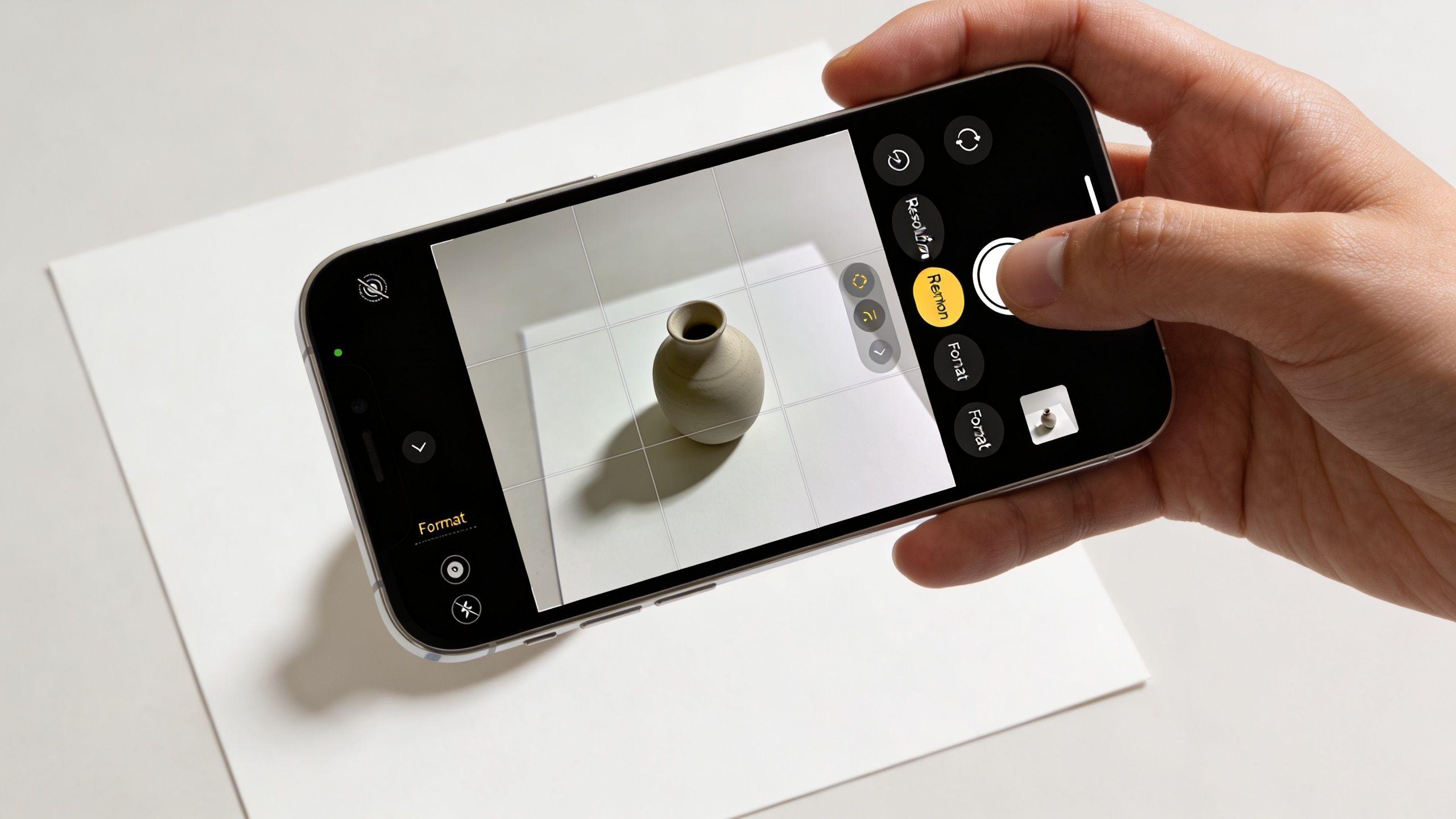 A hand holding an iPhone to photograph a small ceramic vase on a white background surface.