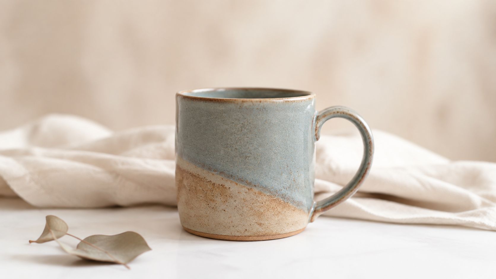 A rustic, two-toned ceramic mug sitting on a marble surface next to a small dried eucalyptus leaf.