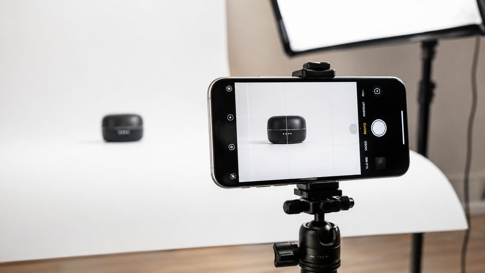 A smartphone on a tripod capturing a product photo of small headphones against a white background.